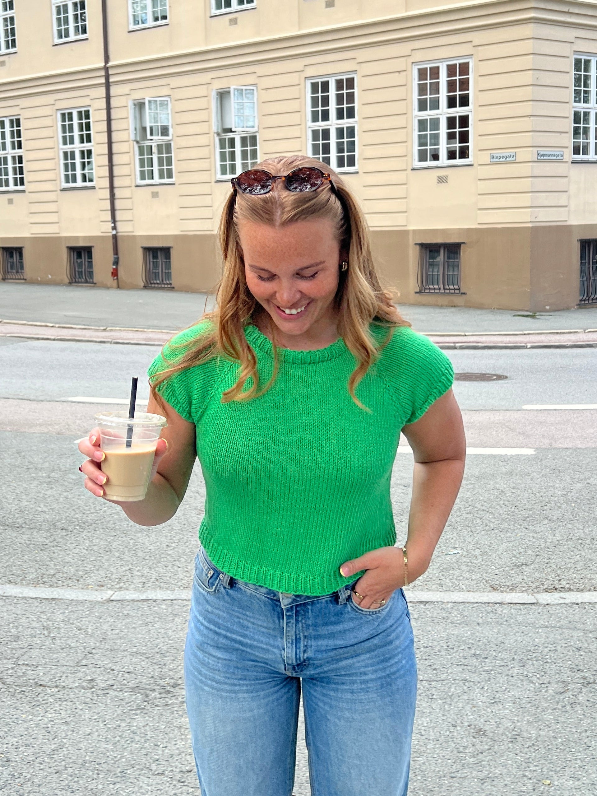Person in a green knitted t-shirt and blue jeans standing on a street with a building in the background