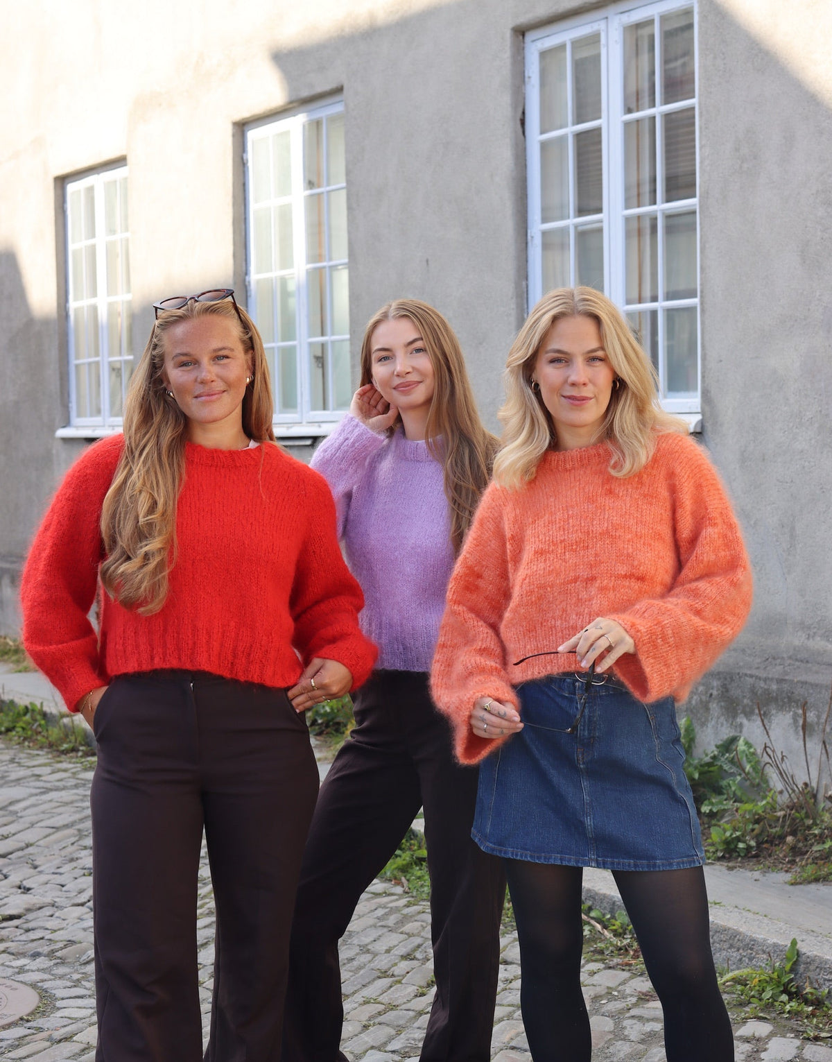 Three women standing outdoors in front of a building, wearing colorful sweaters.